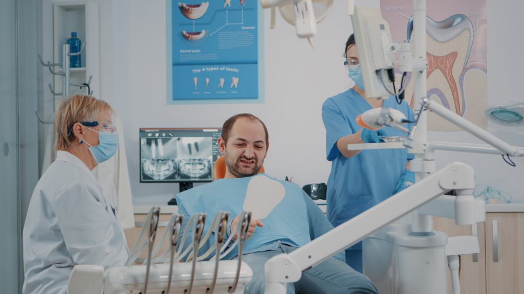 מטופל במרפאת שיניים מחזיק מראה ובוחן את יישור השיניים לאחר טיפול והשתלה
